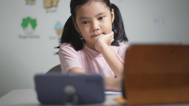 Asian child student study online on computer tablet or kid girl write and learn from home school by video call studying with smartphone or person enjoy learning vocabulary english words in classroom