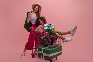 Happy young interracial girlfriends rejoice at gifts bought for holiday. Brunette sits in cart with...