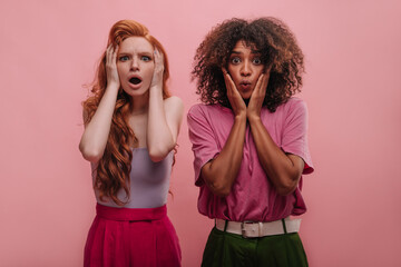 Shocked young european redhead and african brunette on pink background. Amazed and surprised women from around world lay their hands on faces, showing emotion. Lifestyle, female beauty concept