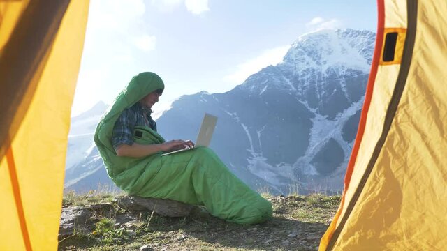 Young man tourist in green sleeping bag opens laptop and types sitting on flat rock against large mountains view from inside tent