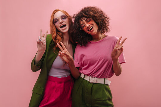 Happy cheerful young girl friends, brunette and redhead show peace sign in empty room. Smiling international women in fuchsia and emerald robes are having fun. Mood, lifestyle, concept