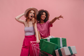Portrait of two interracial young teen girls on pink background. Surprised blue-eyed girl with open mouth holds hat on head next to her friend. She holds out hand with smile to show newly chosen gift