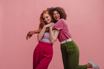Brunette with afro hair hugs embarrassed girlfriend with red hair on pink background. Beauties in...