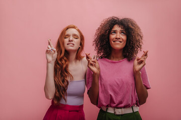 International young women crossed their fingers while biting lip and making wish on pink background. Caucasian slender girl with long red hair posing with african lady dark curls in loose pink T-shirt