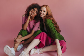 International friendship of two young women caught on camera in bright pink studio. Teenage models from childhood share green trouser suit combined with sports shoes and sit hugging each other. - obrazy, fototapety, plakaty