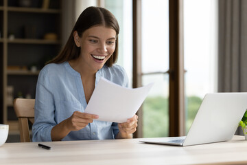 Shocked happy student girl receiving admission letter from university, college, holding, reading paper document at laptop, smiling, laughing, shouting for joy, feeling excited, surprised. Good news - obrazy, fototapety, plakaty
