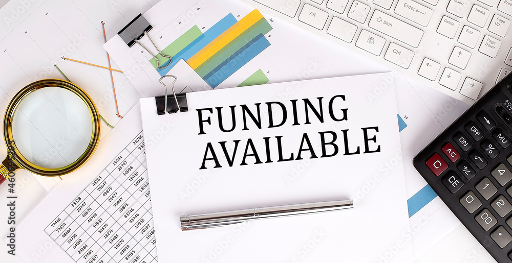 Poster funding available text on the white paper on the light background with charts paper ,keyboard and ca