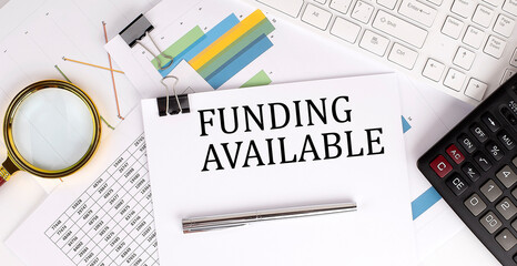 FUNDING AVAILABLE text on the white paper on the light background with charts paper ,keyboard and calculator