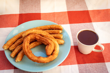 Plate with churros next to a cup of chocolate on a checkered tablecloth.