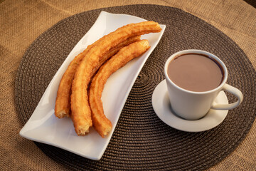 Plate with churros next to a cup of chocolate on a checkered tablecloth.