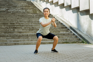Happy Asian lady in comfortable sportswear does squats at training near empty stone steps and concrete wall with decoration