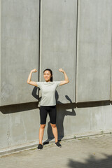 Fototapeta na wymiar Happy brunette Asian sportswoman in light t-shirt shows arm muscles standing by wall with concrete plates on street
