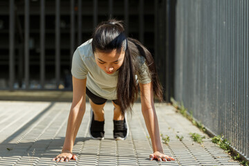 Attractive Asian lady in light t-shirt stands in plank posture on pavement near handrail at outdoor training on city bridge