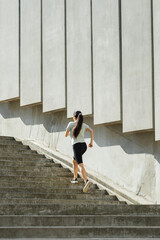 Slim woman with flying hair in tracksuit runs up large stone steps past wall with concrete plates on city street backside view