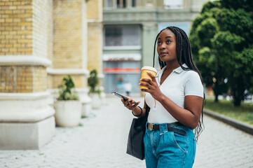 Fototapeta premium Beautiful african american woman using a smartphone and drinking coffee takeaway