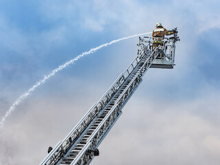 Firefighter pours water on fire. Rescue services during work. Extinguishing fire from hydrant. Firefighter on retractable ladder. Firefighter works on boom of fire engine. Fireman on sky background