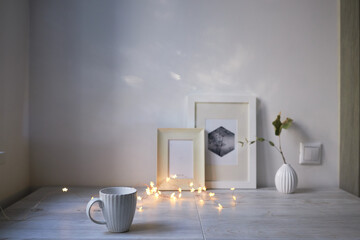 Scandinavian style. Interior Design. A white cup, cozy lights of a luminous garland, a small vase with a dried eucalyptus branch, a photo frame is on the table. Empty space for text