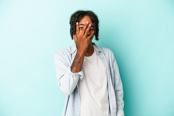 Young african american man isolated on blue background blink at the camera through fingers, embarrassed covering face.