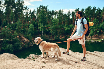 A male hiker with a backpack and a labrador dog stands on a rocky peak, looking at the river and forest. Camping, travel, hiking.
