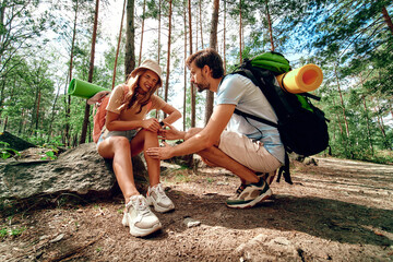 A pair of hikers with backpacks in the woods. The woman bruised her leg, felt pain and sat down on a stone. Camping, travel, hiking. - obrazy, fototapety, plakaty