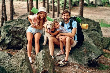 Happy family Mom and dad with their daughter with backpacks and labrador dog sitting on a stone in the forest. Camping, travel, hiking.