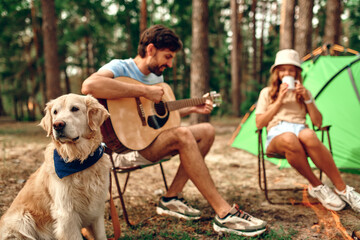 A loving young couple sit near a campfire and a tent, drink coffee, play the guitar with a labrador...