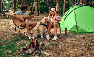 A young family with a child and a labrador dog sit by the campfire near the tent, play the guitar...