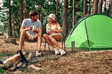 A loving young couple went on a weekend for a picnic, sit near a campfire and a tent, drink coffee...