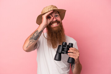 Young caucasian ginger man with long beard holding binoculars isolated on pink background excited keeping ok gesture on eye.