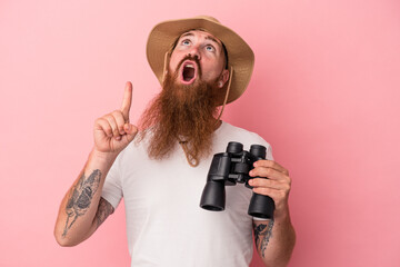 Young caucasian ginger man with long beard holding binoculars isolated on pink background pointing upside with opened mouth.