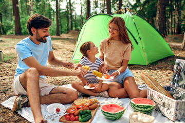 Happy family with a child on a picnic sitting on a blanket near the tent and eating food fried at...
