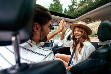 A young couple are giving five to each other in a new car. A man driving a car with his girlfriend and having fun. Buying and renting a car. Travel, tourism, recreation.