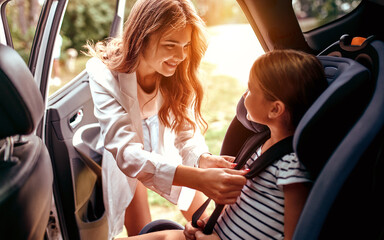 A cute woman mother put her daughter in a car seat and fastens her seat belts. Protection during the trip in the car.