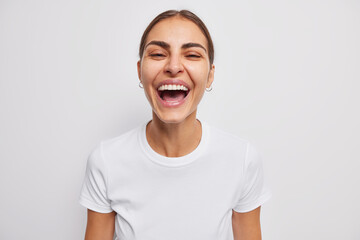 Overjoyed young woman laughs sincerely expresses positive emotions dressed in casual t shirt isolated over white background smiles toothily has clean healthy skin foolishes around chuckles at camera