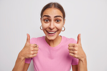 Positive feedback. Glad brunette young European woman keeps thumbs up approves or praises something recommends product smiles happily dressed casually isolated over white background. I like it