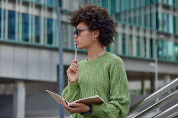 Horizontal shot of thoughtful curly haired Afro American woman holds pen and opened diary makes notes schedule what to do wears sunglasses green jumper poses outside against modern building.