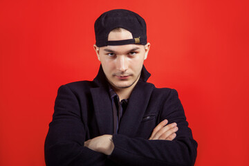 Obraz na płótnie Canvas Portrait of young handsome caucasian man crossing the hands with confident ironic look right to the camera. Black cap and wear, emotional face expression. Red background, copy space.