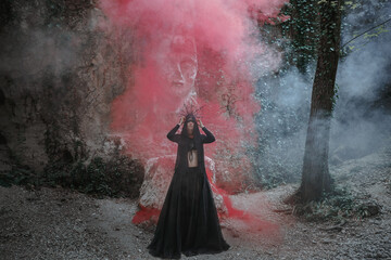 Young woman in witch stands in black dress and crown on her head in dark and foggy forest. - 460614059