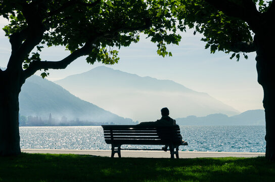 Person in silhouette sitting on his back on a bench looking at a calm lake in the morning with mountains and fog in the background.