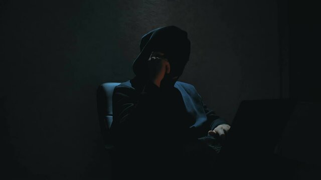 An anonymous hacker in a black hoodie calls his victim on the phone and threatens to crack the database passwords. An Internet hacker in a black balaclava sits in a chair and steals data from a laptop