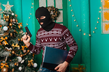 A masked bandit with a pistol stole a laptop for the New Year and escapes. A man in a balaclava holds a computer. Christmas background