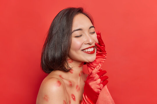 Sideways shot of cheerful young brunette Asian woman keeps eyes closed smiles pleasantly shows white even teeth touches face gently poses with naked shoulders against red background feels happy