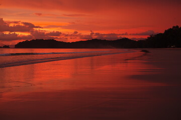 Roter Sonnenuntergang an langem Sandstrand mit flachen Wellen und Reflexionen Sillhouette