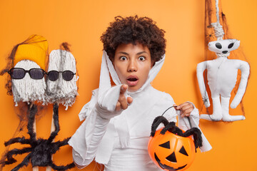 Shocked young African American woman stares at camera points directly at camera with frightened expression holds halloween attributes poses against orange background. Omg what spooky creature
