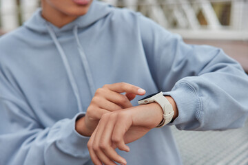 Unrecognizable woman checks results of fitness training on smartwatch uses modern gadget dressed in blue hoodie poses outside looks at display monitors heart rate and pulse leads healthy lifestyle