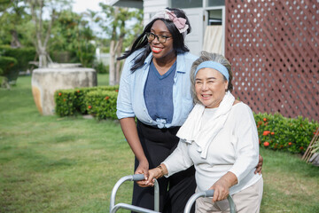 asian senior mother walk with walker and african american carer support in garden at home. old...