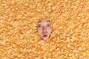 Stunned ethic woman stares bugged eyes has frightened expression holds breath ears dry snack for breakfast drowned in cornflakes makes creative photo from food ingredients. Nutrition concept