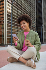 Vertical shot of relaxed curly haired woman sits crossed legs takes rest holds mobile phone checks...