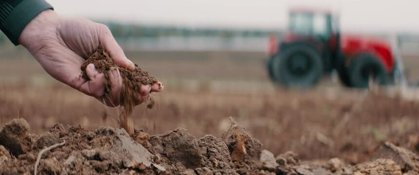 CU on hands, Caucasian male farmer picking up soil, tractor in the background. Shot with 2x anamorphic lens