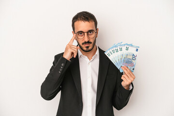 Young caucasian business man holding banknotes isolated on white background pointing temple with finger, thinking, focused on a task.